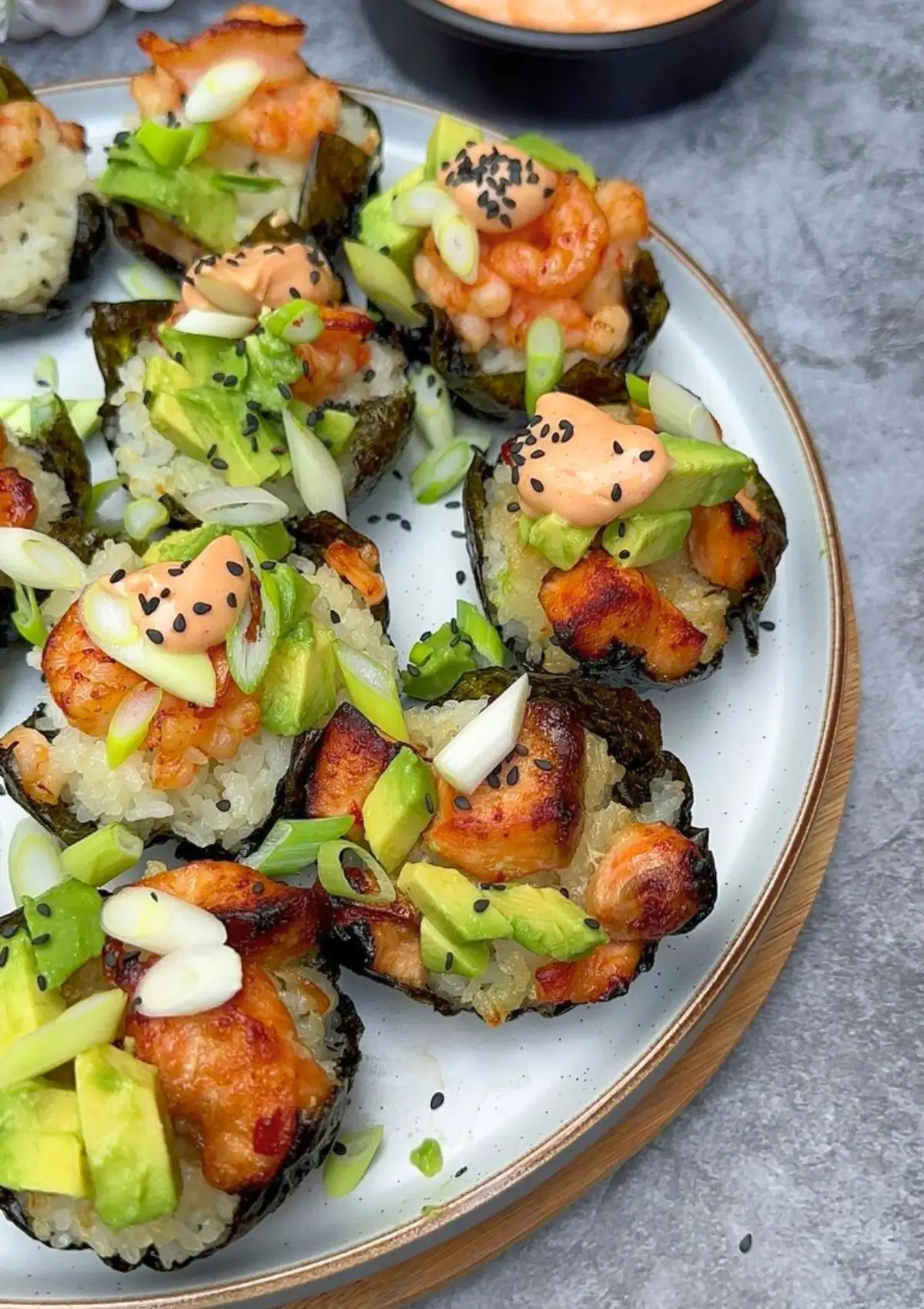 A close-up of baked salmon sushi cups, featuring crispy rice cups filled with tender baked salmon, topped with a mix of shrimp and salmon pieces. Drizzled with sriracha mayo, garnished with black sesame seeds, avocado cubes, and thinly sliced green onions, the sushi cups are beautifully arranged on a platter.