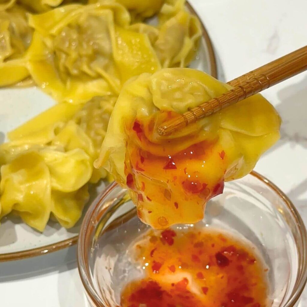 A single wonton dipped in chili sauce held delicately between wooden chopsticks, showing off its golden wrapper with a hint of spicy sauce dripping from the edge.






