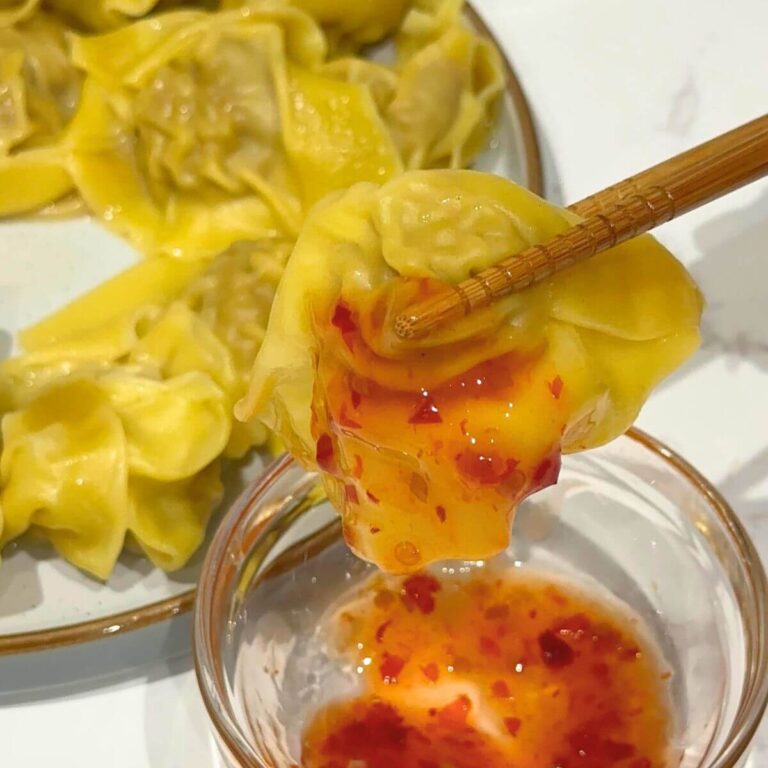 A single wonton dipped in chili sauce held delicately between wooden chopsticks, showing off its golden wrapper with a hint of spicy sauce dripping from the edge.