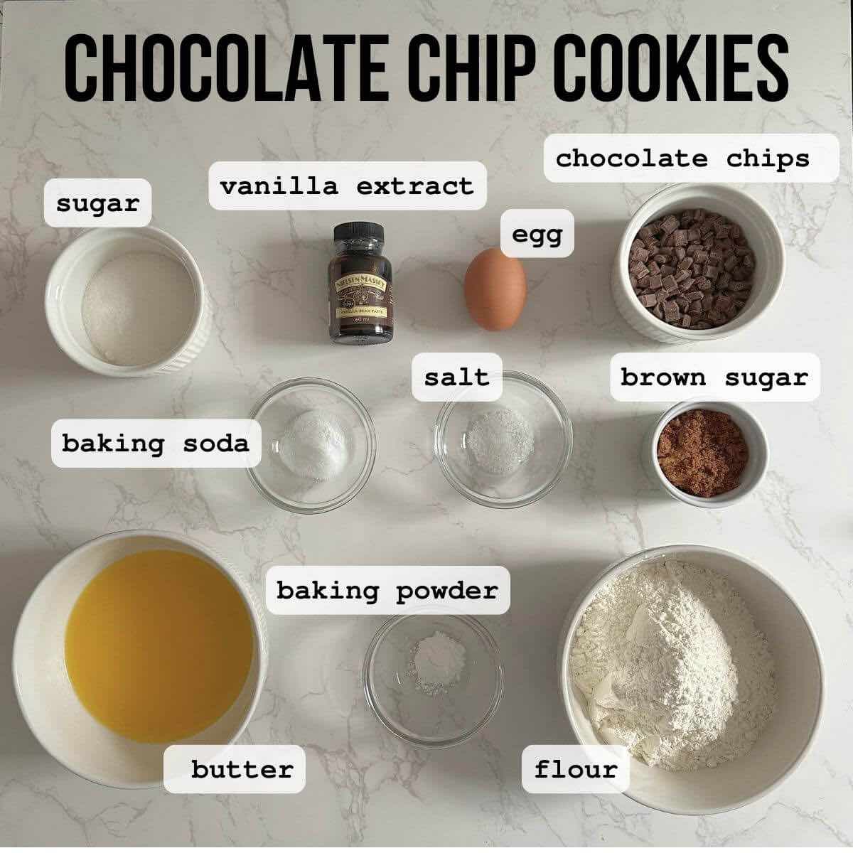 A selection of ingredients for making chocolate chip cookies, including all-purpose flour, baking soda, baking powder, kosher salt, melted unsalted butter, light brown sugar, granulated sugar, vanilla extract, a large egg, semisweet chocolate chips, and flaky sea salt, arranged on a surface.