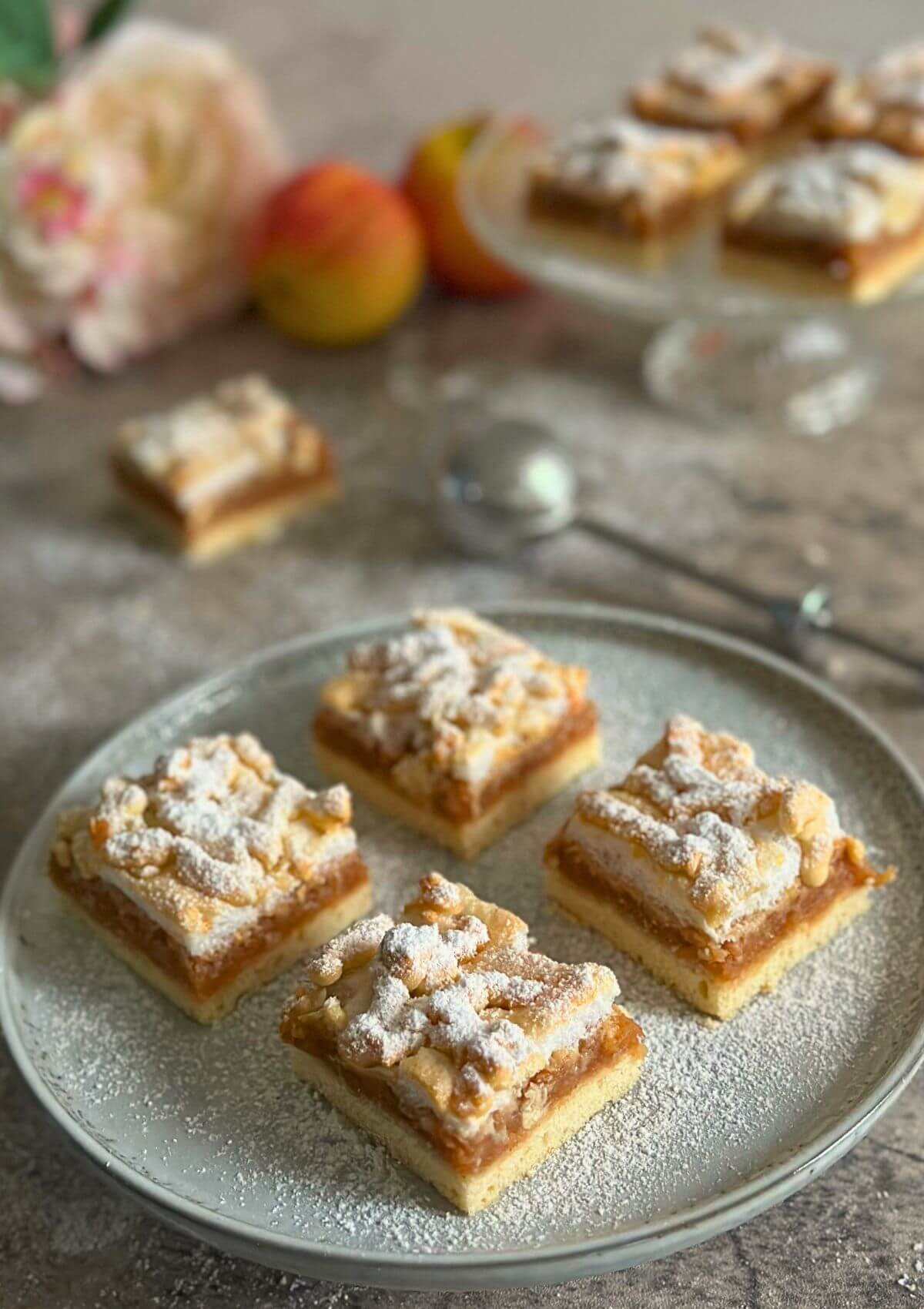 Four apple crumble bars neatly arranged on a plate, with more bars scattered in the background.