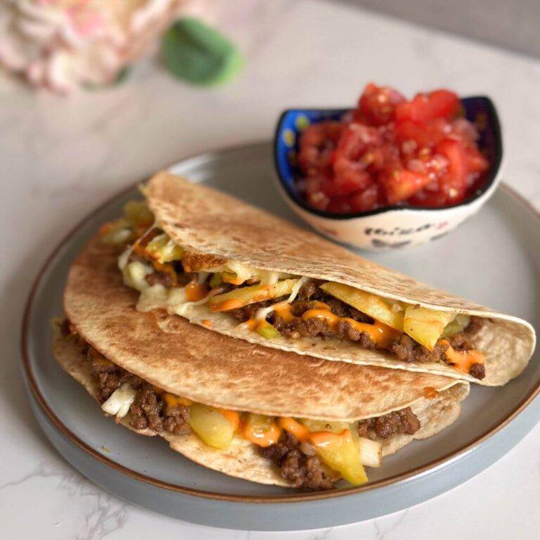 Two tacos filled with seasoned ground beef and crispy fried potatoes, topped with Sriracha mayo and green onions, served alongside a fresh tomato and red onion salad, highlighting vibrant colors and textures.