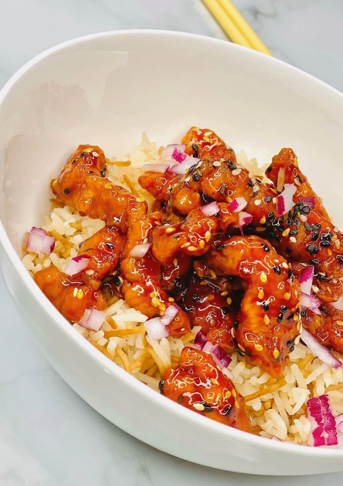 Asian-Style Crispy Sesame Chicken with a sticky sauce served over rice, garnished with red onion chunks