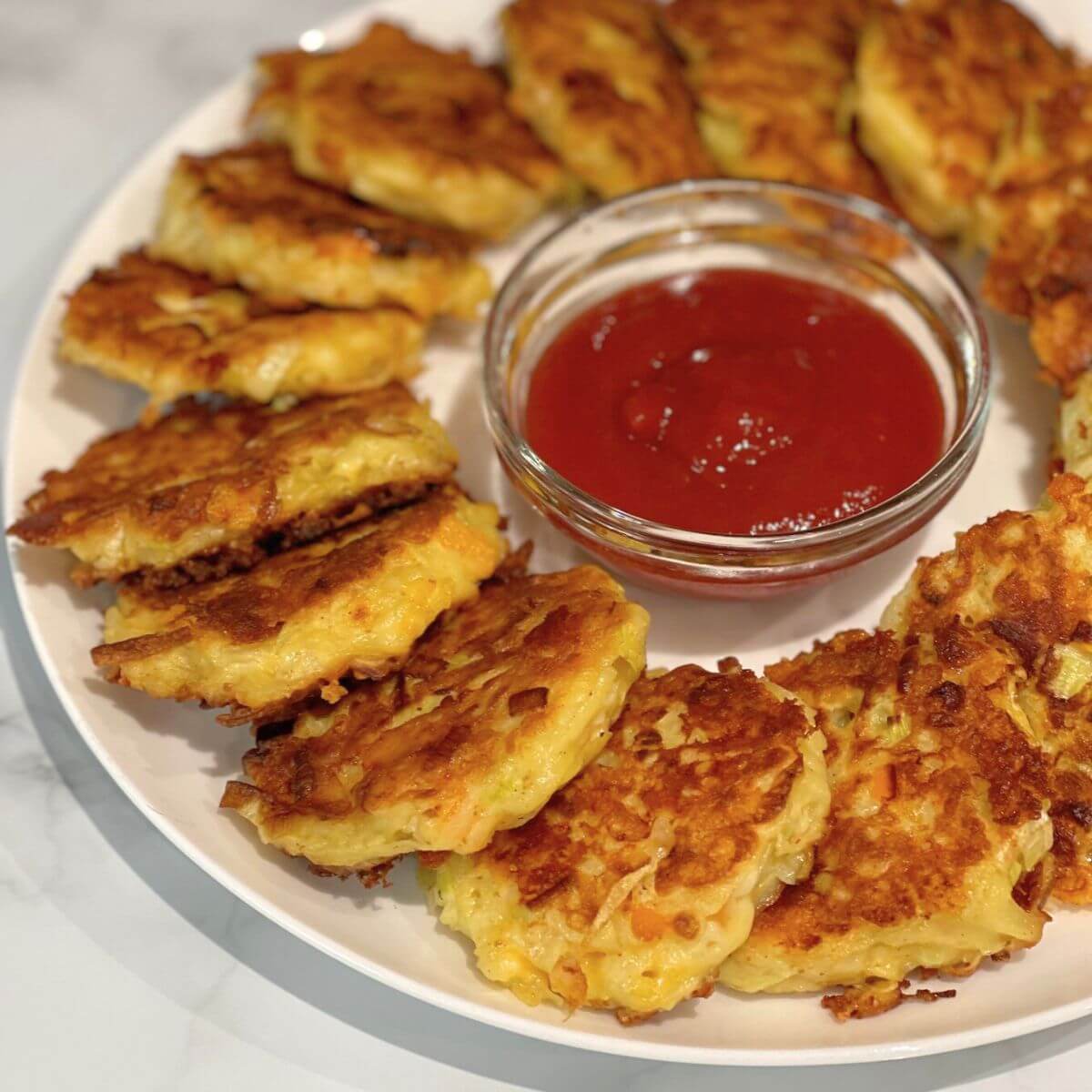 Golden, crispy cheesy leek and potato patties on a white plate, served with a side of ketchup for dipping.