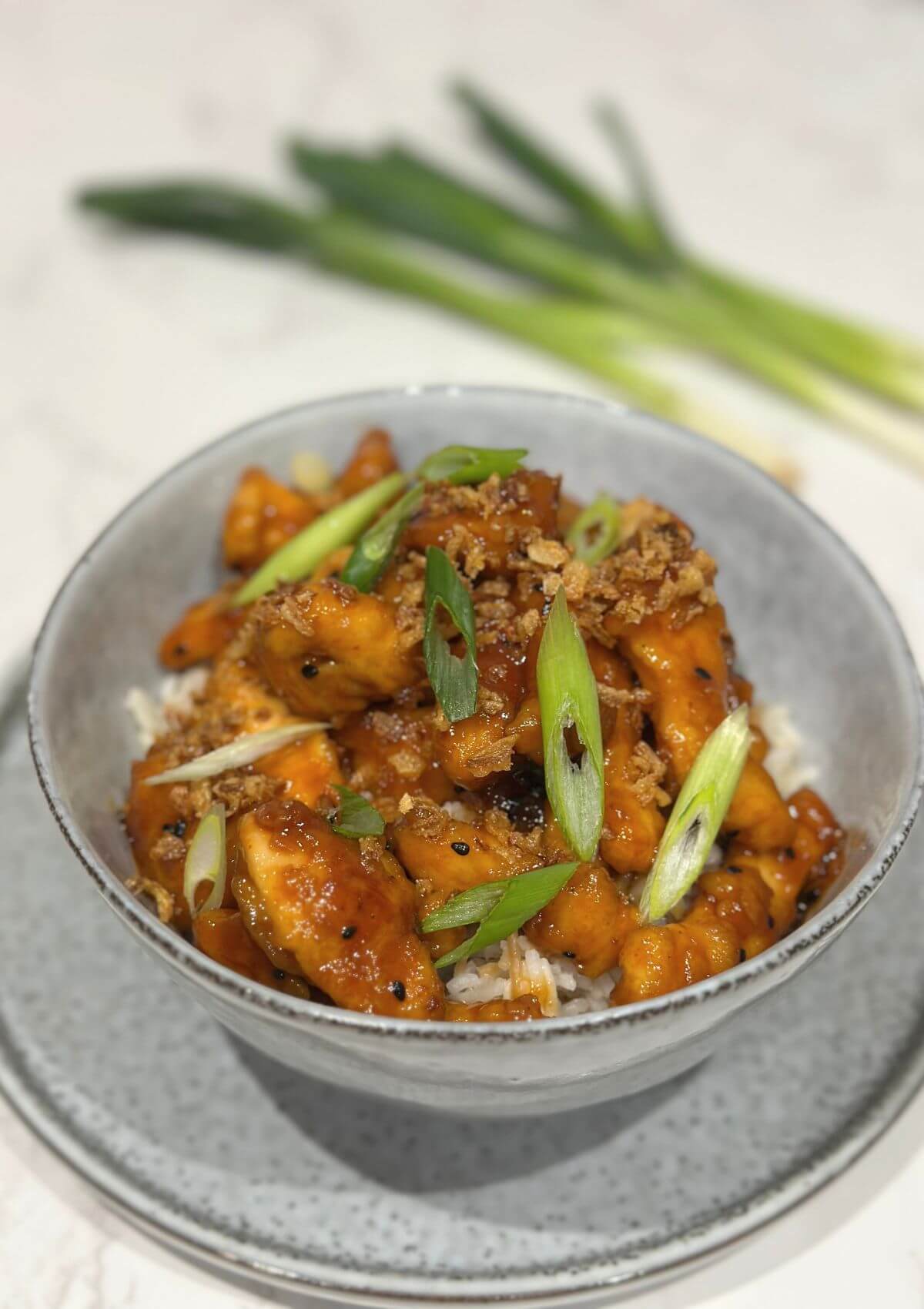 Crispy Sesame Chicken with an Asian Twist in a grey bowl served with rice, onion flakes and green onion cuttings