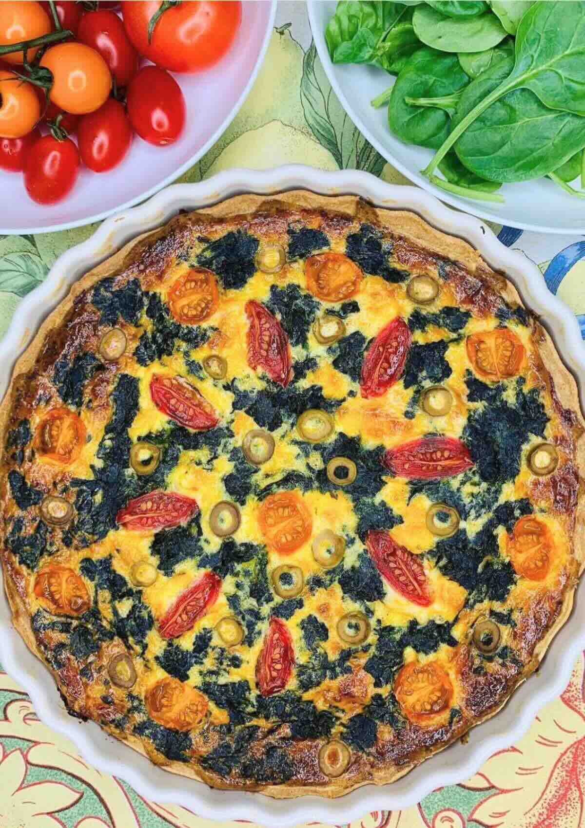 Spinach cherry tomato quiche in a white ceramic dish, surrounded by fresh spinach leaves and cherry tomatoes in small bowls nearby.