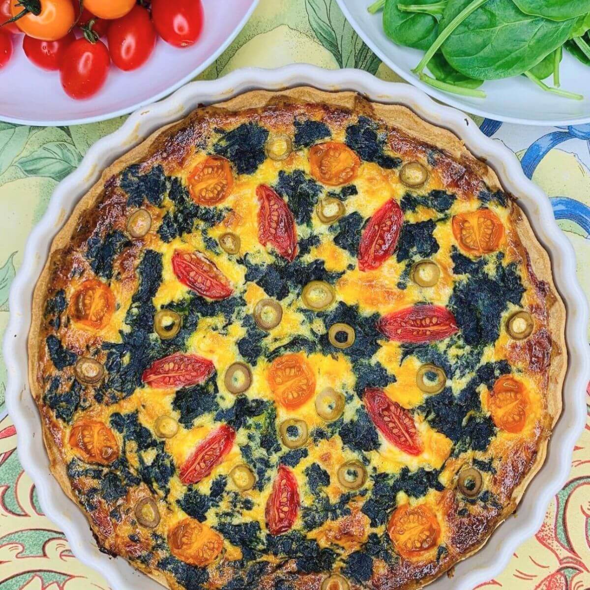 Spinach cherry tomato quiche in a white ceramic dish, surrounded by fresh spinach leaves and cherry tomatoes in small bowls nearby.