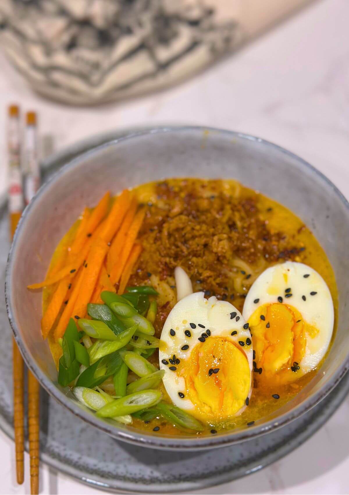 Miso noodle soup with boiled egg, onion flakes, green onions, carrot strings, sesame seeds, and chilli oil.