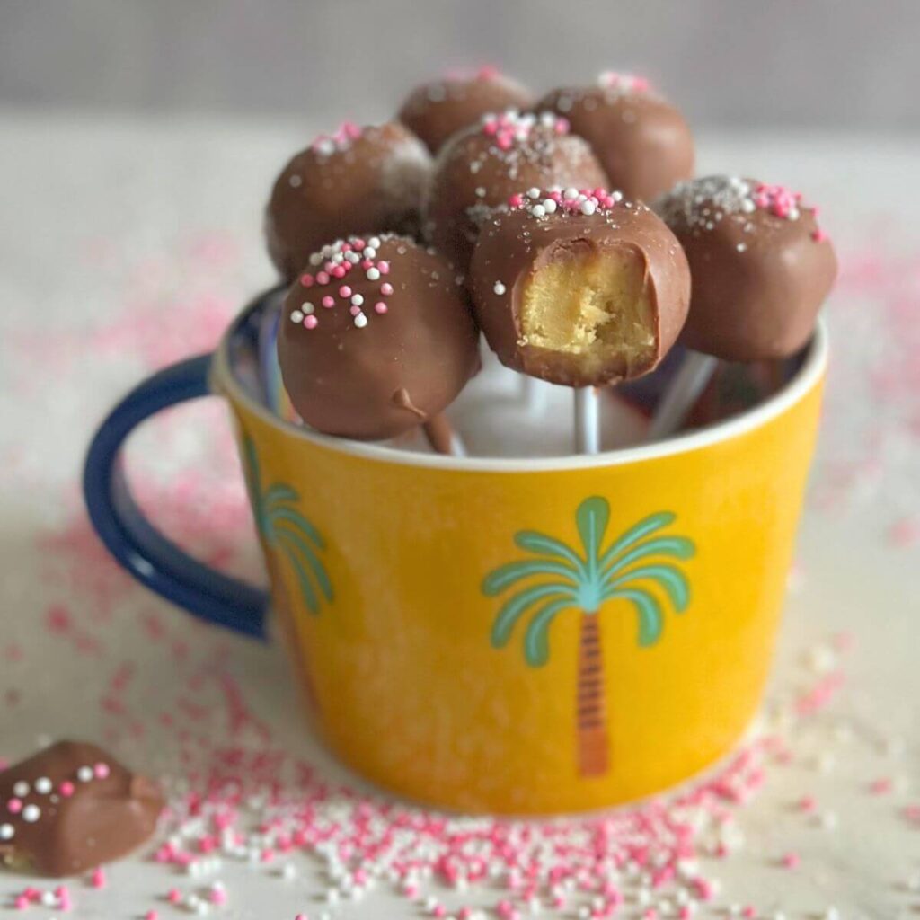 Pistachio Cake Pops in a yellow cup surrounded by sprinkles, one cake pop has a bit take out to see the interior