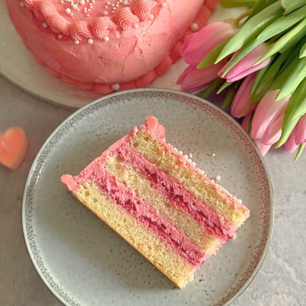 A slice of Raspberry Heart Shaped Cake on a grey plate with some pink tulips in the background and a part of the whole cake