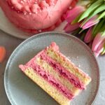 A slice of Raspberry Heart Shaped Cake on a grey plate with some pink tulips in the background and a part of the whole cake