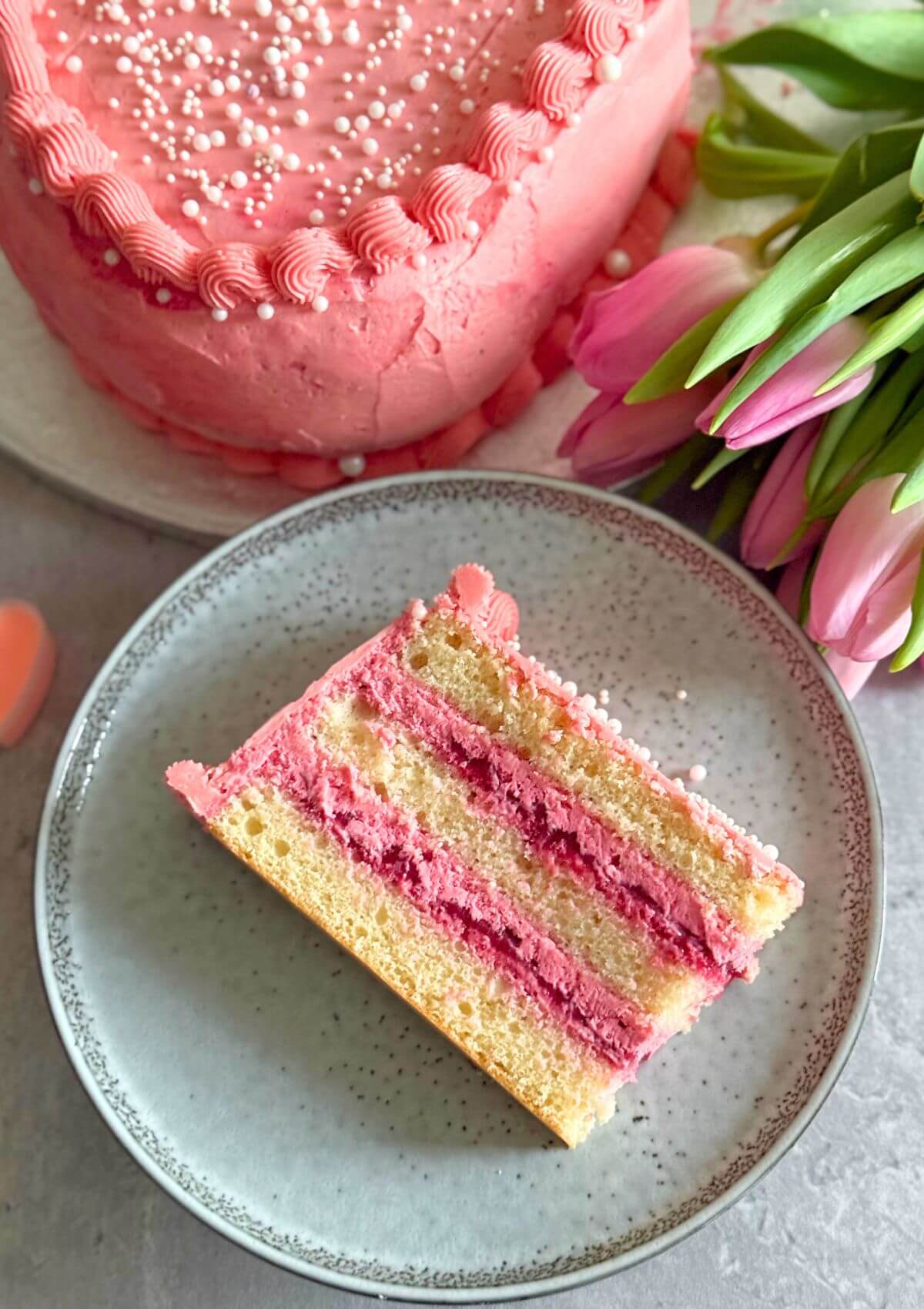 A slice of Raspberry Heart Shaped Cake on a grey plate with some pink tulips in the background and a part of the whole cake